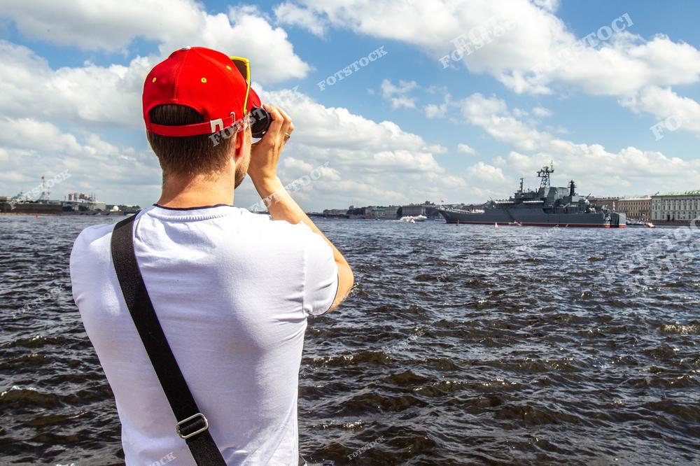 На фотографии изображён турист, снимающий боевые корабли, стоящие на Неве. Мероприятие, скорее всего, связано с празднованием Дня Военно-Морского Флота в Санкт-Петербурге. На заднем плане видны городские здания и часть военно-морской техники. Ясное летнее небо и водная гладь создают праздничную атмосферу.